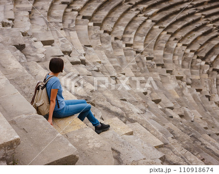 Woman tourist sitting in amphitheater enjoying solitude. travel to ancient city of Side, Antalya coast of Turkey in tourist low season. view of ancient theatre or Amphitheatre in Side, Turkey. Woman tourist sitting in amphitheater enjoying solitude. travel to ancient city of Side, Antalya coast of Turkey in tourist low season. view of ancient theatre or Amphitheatre in Side, Turkey. 110918674