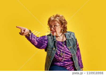 Elderly woman with cigarette in mouth expressive pointing somewhere dressed denim jacket and tie-dye T-shirt. Directive emotions. Elderly woman with cigarette in mouth expressive pointing somewhere dressed denim jacket and tie-dye T-shirt. Directive emotions. 110918998
