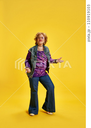 Elderly emotional woman in denim jeans purple top dancing against vibrant yellow background. 110919103