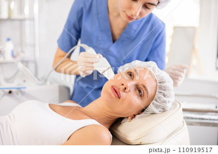 Young female client experiencing facial skin peeling using ultrasonic scrubber 110919517