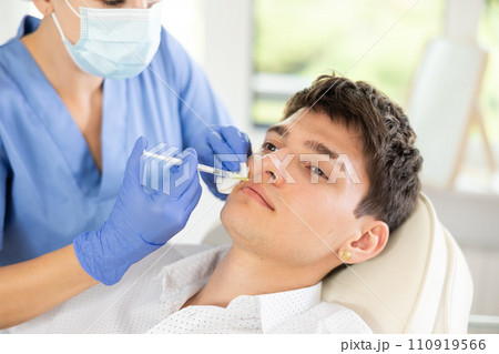 Closeup face of young male client receiving injections during lip enhancement procedure, professional cosmetologist hands in gloves holding syringe 110919566