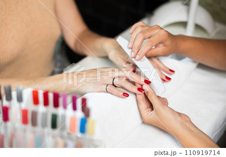 Hands of young girl manicurist perform trimming saws nails to elderly lady client. 110919714