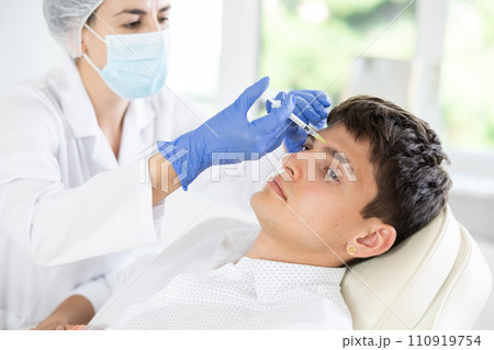 Professional cosmetologist hands in gloves performing lips enhancement treatment to male client, making injection of lip fillers 110919754
