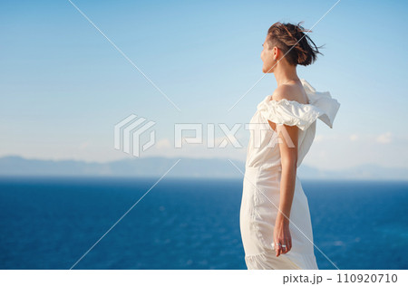 Beautiful Asian young woman in white dress outdoor in flower field under Rhodes city above sea during sunset. embracing fresh air and engaging in outdoor activities. strong wind - element of freedom 110920710