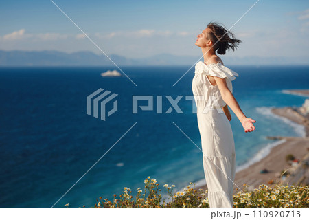 Beautiful Asian young woman in white dress outdoor in flower field under Rhodes city above sea during sunset. embracing fresh air and engaging in outdoor activities. strong wind - element of freedom Beautiful Asian young woman in white dress outdoor in flower field under Rhodes city above sea during sunset. embracing fresh air and engaging in outdoor activities. strong wind - element of freedom 110920713