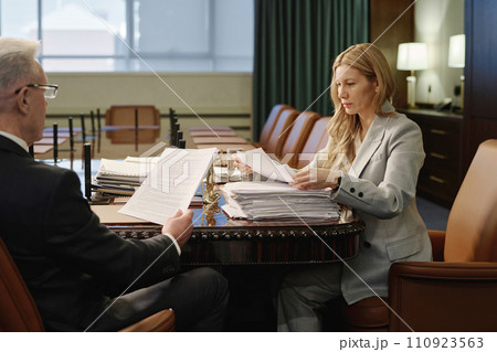 Female lawyer and her senior colleague sitting at wooden desk in boardroom and reading documents Female lawyer and her senior colleague sitting at wooden desk in boardroom and reading documents 110923563