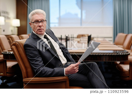 Senior businessman posing in armchair with documents in his hands and confidently looking at camera Senior businessman posing in armchair with documents in his hands and confidently looking at camera 110923596