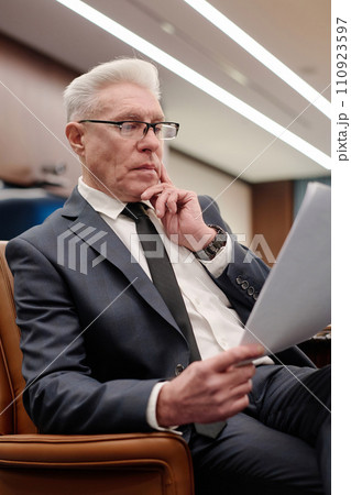 Senior businessman in formalwear sitting in armchair and thoughtfully reading information on paper 110923597