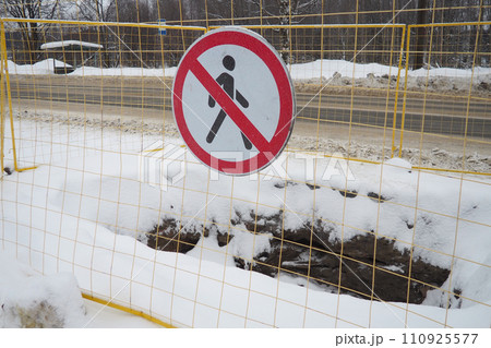 No passage sign, do not pass. Man crossed out with a red line. Round prohibitory road sign. Forbidden. A hole dug in the ground, surrounded by a metal fence. Winter, snow and snowdrifts near the road No passage sign, do not pass. Man crossed out with a red line. Round prohibitory road sign. Forbidden. A hole dug in the ground, surrounded by a metal fence. Winter, snow and snowdrifts near the road 110925577