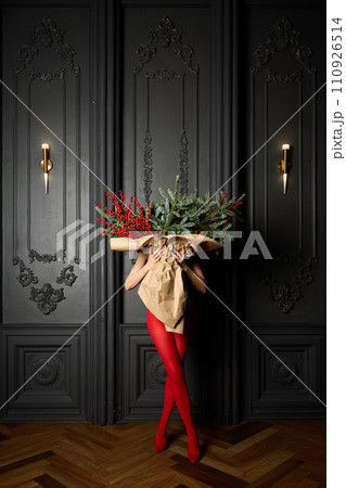 A long girl in red tights and high-heeled shoes holds a huge bouquet of fir branches and red berries wrapped in cardboard paper, she closes with a bouquet, photo studio with grey wall background 110926514