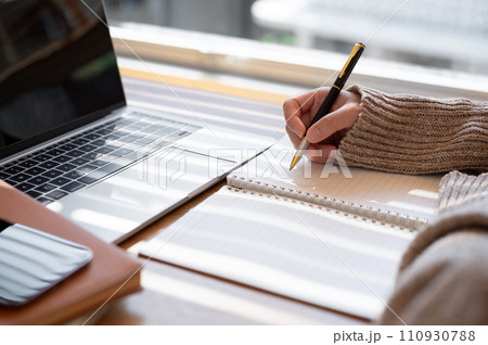 Close-up image of a woman is writing something in her notebook and working on her laptop indoors. 110930788