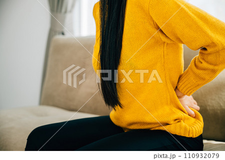 Portraying backache, Asian woman on sofa suffers from unbearable lower back pain. Illustrating chronic back pain discomfort and necessity of medical care for back problems. health care concept 110932079