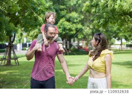 A man and woman are walking together holding hands in a beautiful green park, while the father holds a giggling small child on his shoulder. A man and woman are walking together holding hands in a beautiful green park, while the father holds a giggling small child on his shoulder. 110932638