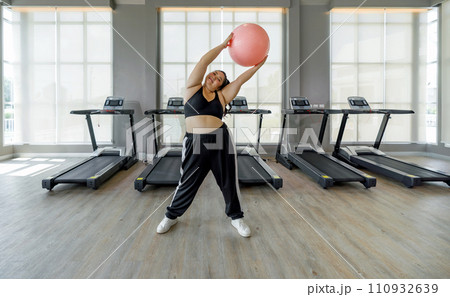 A woman is in a gym, gripping a bright pink ball with both hands, ready to exercise. 110932639