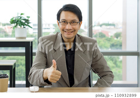 A man wearing a formal suit smiles and shows a thumbs up sign with his hand to express approval. 110932899