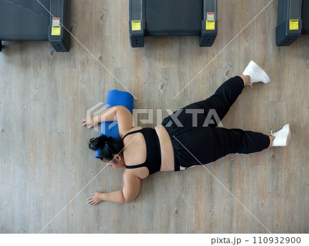 A plus-size woman lies on a gym floor, resting or unmotivated, surrounded by exercise equipment. A plus-size woman lies on a gym floor, resting or unmotivated, surrounded by exercise equipment. 110932900
