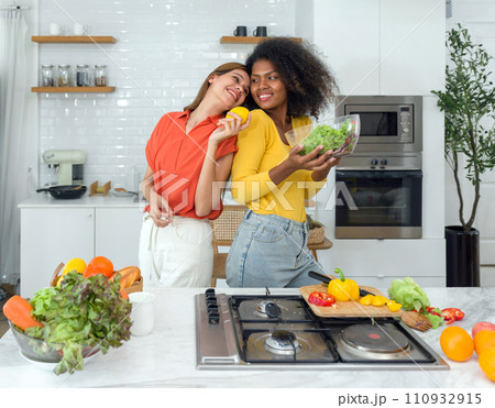 Two women stand in a kitchen holding various fruits and vegetables, smiling. 110932915
