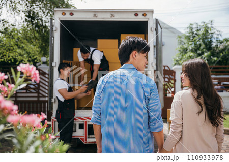 With professional help a couple moves into their new house smoothly. The delivery team collaborates unloading and lifting cardboard boxes for an efficient relocation. Moving Day Concept With professional help a couple moves into their new house smoothly. The delivery team collaborates unloading and lifting cardboard boxes for an efficient relocation. Moving Day Concept 110933978