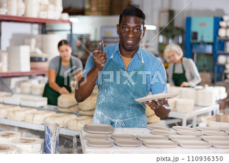 Portrait of african american worker cutting burrs off freshly made earthenware cups and plates with knife 110936350