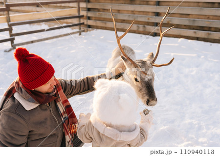 Back view of little child boy and loving father feeding cute young reindeer on snowy farm on sunny winter day. Concept of ecotourism at wintertime. 110940118