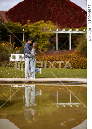 A couple in love hugs on the shore of a city pond in the European town. love story against the backdrop of autumn nature. Embraced by Love: Couple by the City Pond in a European Town. Autumn Affair 110944677