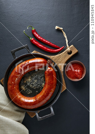 Ring of fried pork sausage on a black cast iron frying pan with hot pepper and ketchup on a black background. Top view with copy space 110945845