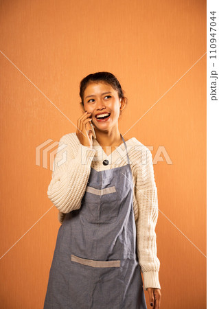 smiling Asian girl wearing apron making cell phone call smiling Asian girl wearing apron making cell phone call 110947044