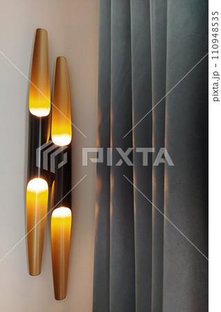 Two modern sleek wall lights casting warm glow beside elegant dark gray curtains Two modern sleek wall lights casting warm glow beside elegant dark gray curtains 110948535
