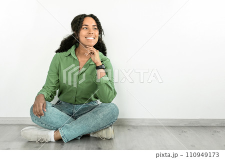 Black woman looks aside with smile, sitting near white wall Black woman looks aside with smile, sitting near white wall 110949173