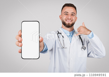 Young cheerful doctor showing phone with blank screen and gesturing call me 110949591