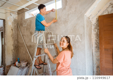 smiling wife holding a ladder as husband measures the height of a vent when building a new house 110949617