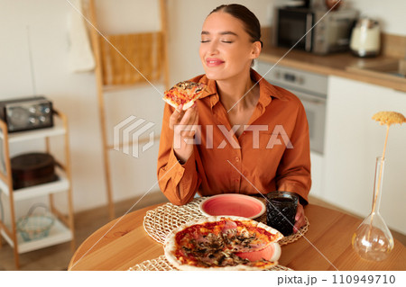 Woman savoring a slice of pizza in a cozy kitchen Woman savoring a slice of pizza in a cozy kitchen 110949710