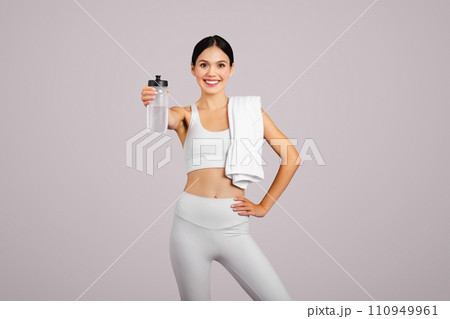 Smiling slim caucasian lady in sportswear with towel showing bottle of water, isolated on grey studio background Smiling slim caucasian lady in sportswear with towel showing bottle of water, isolated on grey studio background 110949961