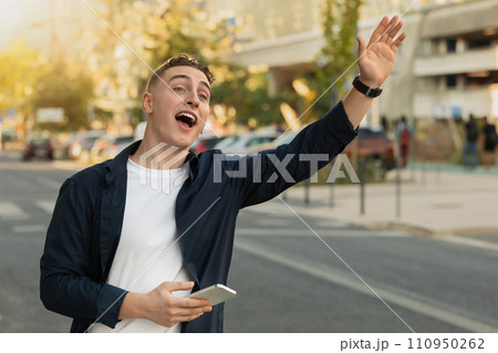 Cheerful handsome millennial caucasian guy in casual, use phone in city street, stop taxi Cheerful handsome millennial caucasian guy in casual, use phone in city street, stop taxi 110950262