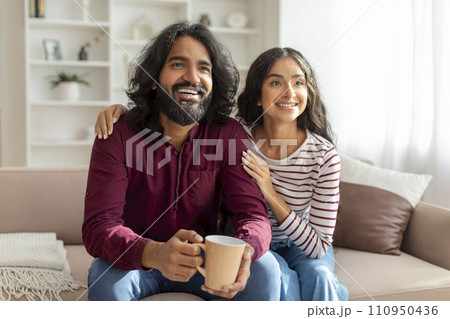 Happy millennial indian man and woman drinking coffee, watching movie 110950436
