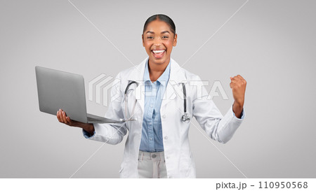 Happy black millennial lady doctor in white coat hold computer, rise fist sign 110950568