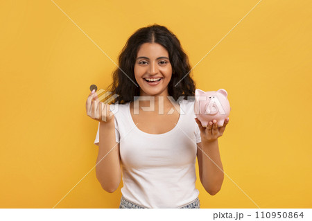 Emotional young woman with piggybank and coin on yellow Emotional young woman with piggybank and coin on yellow 110950864