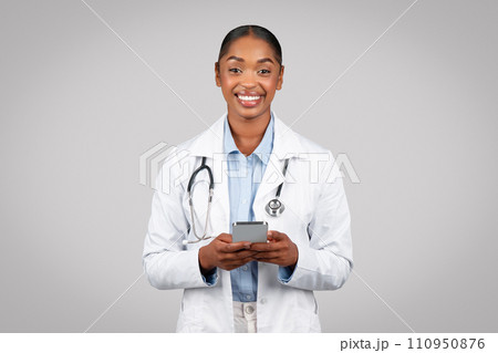 Positive millennial black lady doctor in white coat, typing on mobile phone, chatting on gadget Positive millennial black lady doctor in white coat, typing on mobile phone, chatting on gadget 110950876