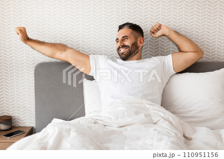man smiling and stretching arms while sitting up in bed man smiling and stretching arms while sitting up in bed 110951156
