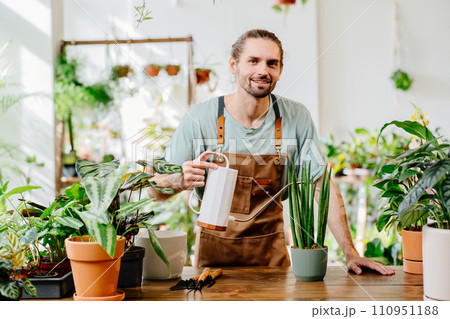 Young man wearing apron carefully watering potted houseplants at home indoors. Concept of plants care and home garden. 110951188