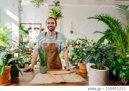 Handsome Caucasian man wearing apron wrapping a potted plant in paper with care in greenery at floral shop. Enjoying his job at cozy flower shop. Urban jungle and plant parent concept. 110951221