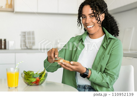 Black lady smiles making sandwich for breakfast sitting at kitchen 110952139