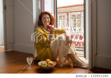 Smiling senior woman sitting by open balcony door and eating ripe apple 110952280
