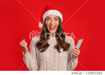 woman in Santa hat celebrates holding phone shaking fist, studio woman in Santa hat celebrates holding phone shaking fist, studio 110952283