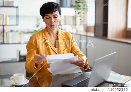 businesswoman reading business papers near her laptop in domestic office 110952336