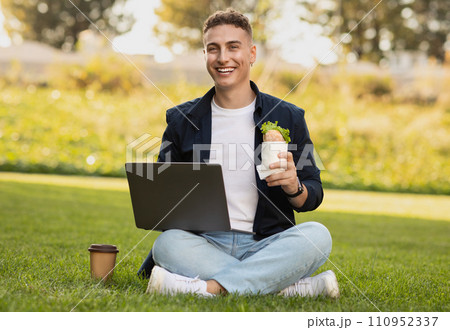 Glad millennial caucasian man in casual enjoy sandwich, work on computer, has lunch in park 110952337