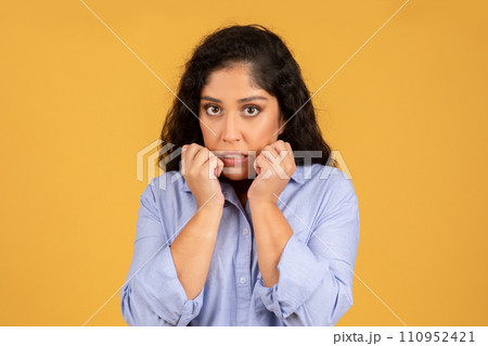A worried young woman with curly hair wearing a blue shirt bites her nails and looks directly at the camera A worried young woman with curly hair wearing a blue shirt bites her nails and looks directly at the camera 110952421