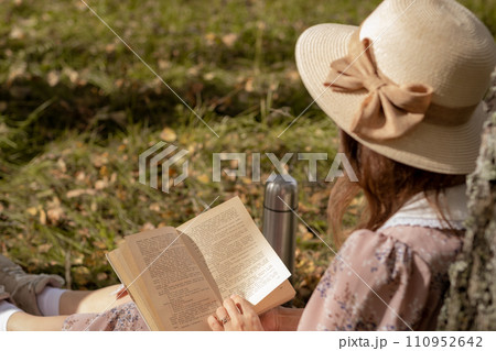 A young beautiful woman in a dress and a round hat reads a book outdoors in the forest and drinks tea. Romantic and vintage photo of a beautiful girl. Reading and relaxation A young beautiful woman in a dress and a round hat reads a book outdoors in the forest and drinks tea. Romantic and vintage photo of a beautiful girl. Reading and relaxation 110952642