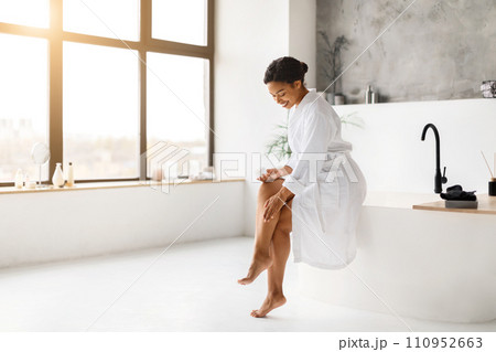 Happy African American Lady Applying Moisturizer Cream On Legs In Bathroom Happy African American Lady Applying Moisturizer Cream On Legs In Bathroom 110952663