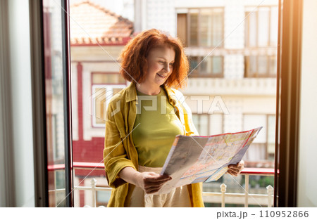 Redhead senior woman checking travel map while standing alone on home balcony 110952866